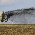 Ein großer Tankanhänger sprüht Flüssigkeit auf ein Feld. Die Reifen des Anhängers sind robust. Der Himmel ist blau und das Feld scheint brach zu liegen.