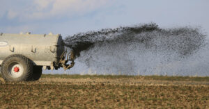 Ein großer Tankanhänger sprüht Flüssigkeit auf ein Feld. Die Reifen des Anhängers sind robust. Der Himmel ist blau und das Feld scheint brach zu liegen.