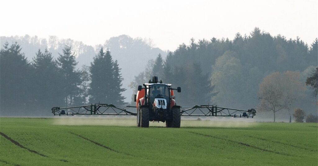 Traktor mit Sprühgestänge verteilt Dünger über grünes Feld, umgeben von Wald am Horizont. Keine Menschen sichtbar.